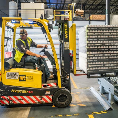 A warehouse worker maneuvers a forklift to transport crates for brewing company storage.